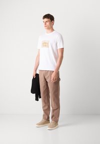 Hvit kortermet t-skjorte med grafisk trykk, kombinert med beige cargobukser og lyse beige sneakers, holder en svart genser.