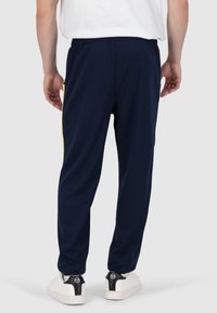Marineblaue Jogginghosen mit strukturiertem Finish, elastischem Bund, Seitentaschen und kontrastierenden gelben Akzenten entlang der Nähte. Weiße Sneakers sind sichtbar.