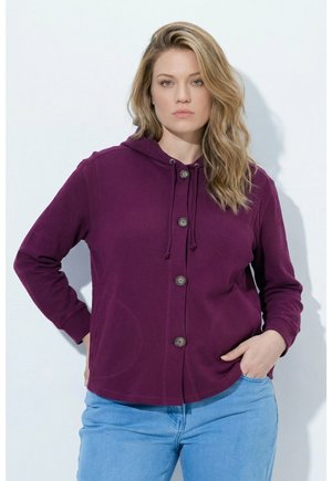 Frau mit langen blonden Haaren, die einen lila Kapuzenpullover mit Knöpfen und hellblaue Jeans trägt, steht mit einer Hand in der Tasche vor einem einfarbigen Hintergrund.