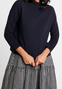 Granatowy sweter o prążkowanej fakturze z wysokim dekoltem, zestawiony z czarno-białą spódnicą w kratę o warstwach. Teksturowany materiał i luźny krój.