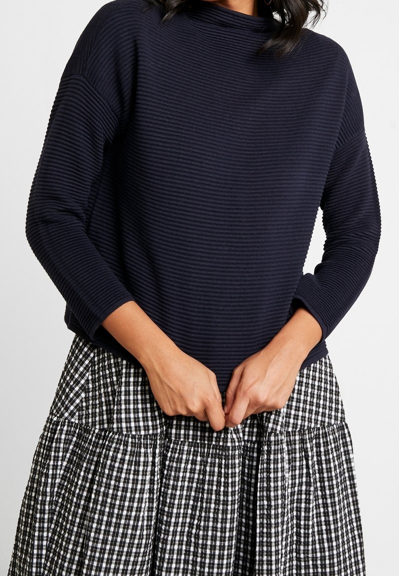 Granatowy sweter o prążkowanej fakturze z wysokim dekoltem, zestawiony z czarno-białą spódnicą w kratę o warstwach. Teksturowany materiał i luźny krój.