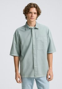 Lichtgroen shirt met korte mouwen, met knoopsluiting aan de voorkant, een borstzak en een relaxte pasvorm. Gemaakt van textiel met een structuur, gecombineerd met lichtblauwe jeans.