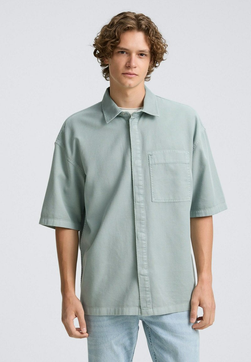 Lichtgroen shirt met korte mouwen, met knoopsluiting aan de voorkant, een borstzak en een relaxte pasvorm. Gemaakt van textiel met een structuur, gecombineerd met lichtblauwe jeans.