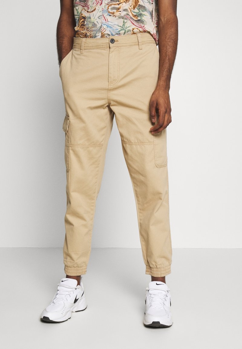 Pantalon cargo beige avec poignets élastiques porté avec des baskets blanches Nike et une chemise à manches courtes à imprimé tropical.