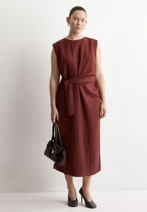 Mujer de pie contra una pared blanca, vistiendo un vestido midi burdeos sin mangas atado a la cintura, sosteniendo un bolso negro y calzando tacones altos negros.