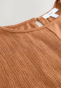 Haut de couleur rouille présentant un tissu texturé à côtes avec une ouverture en forme de clé à l'arrière. Fabriqué en matériau léger avec une finition douce.