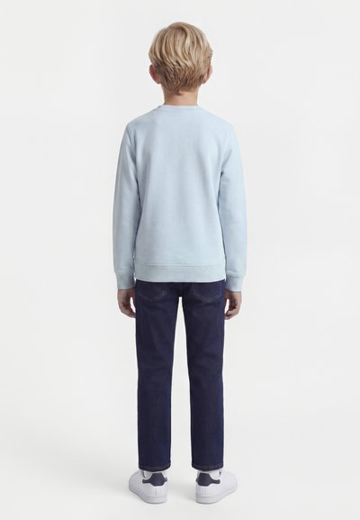 Blondes Kind, von hinten gesehen, trägt ein hellblaues Langarmshirt, dunkelblaue Jeans und weiße Turnschuhe vor einem schlichten weißen Hintergrund.