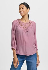 Lys pink bluse med blomsterlignende læg ved halsen, trekvartærmer med flæser og en afslappet pasform.