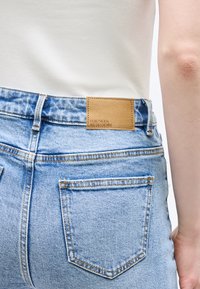 Κοντινή λήψη του πίσω μέρους ανοιχτόχρωμων μπλε τζιν με δερμάτινο λογότυπο σε καφέ απόχρωση και λευκή ριμπ μπλούζα που είναι μισο tucked μέσα.