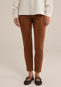 Žena nosící hnědé úzké semišové kalhoty s černými síťovanými Mary Jane botami a krémovou manžestrovou košili, stojící na béžové podlaze.