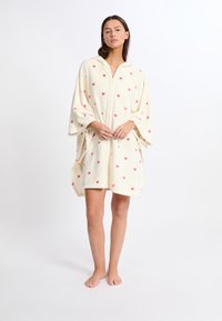 Asciugamano con cappuccio in fleece crema con pattern a cuori rossi sparsi. Presenta maniche larghe e una zip frontale per una facile vestibilità. Texture morbida.