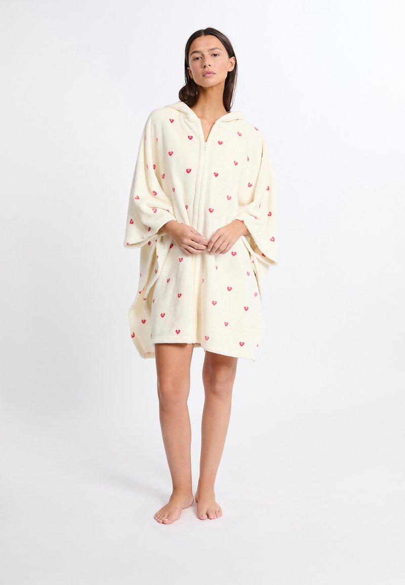 Asciugamano con cappuccio in fleece crema con pattern a cuori rossi sparsi. Presenta maniche larghe e una zip frontale per una facile vestibilità. Texture morbida.