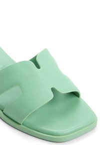 Sandale glissante vert clair avec une large bride à design découpé, texture lisse et bout carré. Semelle plate avec un minimum de détails.