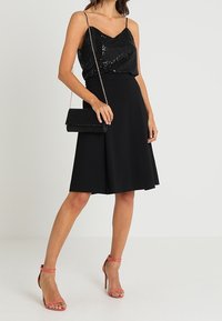 Kvinna som bär en svart paljettlinnen, svart knälång kjol, rosa högklackade sandaler och håller en svart kuvertväska med kedjesnodd.