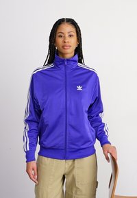 Giacca a zip viola realizzata in materiale lucido, con strisce bianche sulle spalle e un logo ricamato sul petto.