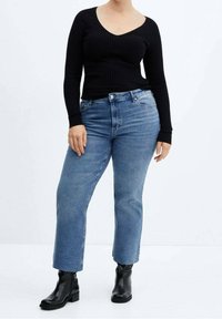 Frau trägt schwarzen, gerippten Langarmpullover, blaue Jeans mit geradem Bein und schwarze Stiefeletten, steht vor weißem Hintergrund.