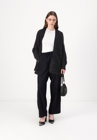Svart fluffy cardigan over en hvit topp, paret med marineblå stripete vide bukser og svarte patent hæler. Tilbehøret inkluderer en liten svart veske.
