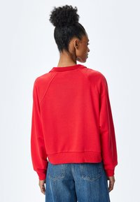 Czerwony sweter z prążkowanym kołnierzem i mankietami, bocznymi szwami, skróconym krojem, noszony na niebieskich dżinsach z wysokim stanem, minimalna widoczna hardware.