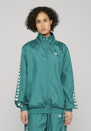 Femme aux cheveux bouclés portant un survêtement zippé bleu sarcelle avec des bandes blanches en forme de chevron sur les manches et le pantalon, debout devant un fond gris uni.