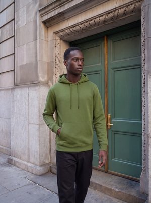 Junger Mann in grünem Hoodie und schwarzen Hosen läuft auf einem Bürgersteig in der Stadt an einer grünen Tür, die in eine steinerne Gebäudefassade eingelassen ist, vorbei.