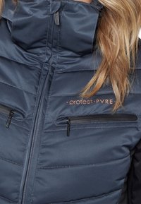 Marineblaue gesteppte Jacke mit schwarzen Reißverschlusstaschen auf der Brust, hohem Kragen und sichtbar hellbraunem Haar auf der rechten Seite.