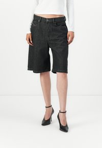Svarte denimshorts med løs passform, oppbrett på kanten og framlommer. Kombinert med svarte ankelrem-hæler. Hvit langermet topp synlig.