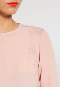 Femme portant un haut rose pâle avec un détail en dentelle florale sur l'épaule, rouge à lèvres rouge vif et peau lisse sur un fond uni.