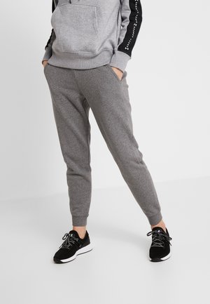 Persoon die grijze joggingbroek en bijpassende hoodie draagt, staat met handen in de zakken, draagt zwart-witte sneakers op een effen achtergrond.