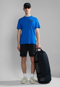 Kék póló, fekete rövidnadrág és fekete sapka. Fekete hengeralakú sporttáska, texturált felülettel és logórészletekkel. Fehér edzőcipő zoknival.