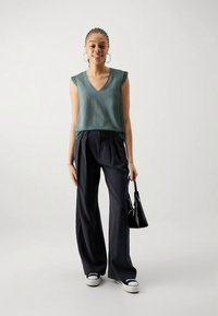 Kvinde med flettet hår iført en grøn ærmeløs top, sorte vide bukser, hvide sneakers og bærer en lille sort håndtaske.