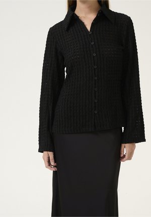 Femme portant une chemise noire texturée à manches longues, boutonnée avec col, et une jupe noire, debout devant un fond uni.