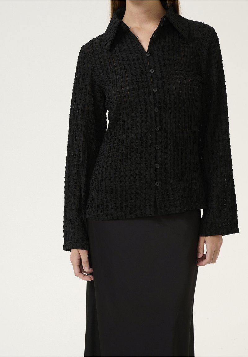 Femme portant une chemise noire texturée à manches longues, boutonnée avec col, et une jupe noire, debout devant un fond uni.
