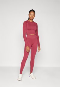 Bordó sportos szett, amely egy testhez simuló, hosszú ujjú crop topot és magas derekú leggingset tartalmaz, sima, rugalmas anyagból. Fehér sportcipőt visel.