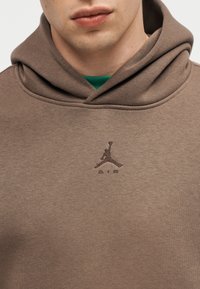 Jeune personne portant un sweat à capuche marron avec le logo Jumpman Air marron foncé brodé sur la poitrine.