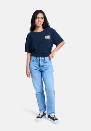 Frau mit langen dunklen Haaren, die ein schwarzes Grafik-T-Shirt, hellblaue Jeans und schwarz-weiße Sneakers trägt, steht mit einer Hand in der Hüfte.