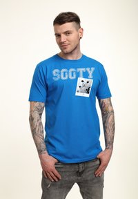 Modré bavlněné tričko s bílým nápisem "SOOTY" a grafikou černobílé kočky v bílém rámečku v oblasti levé hrudi.