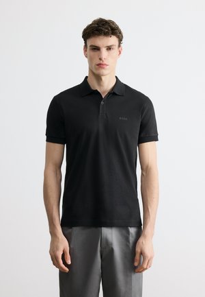 Νεαρός άνδρας που φοράει μαύρο πόλο μπλουζάκι BOSS και γκρι παντελόνι, στέκεται μπροστά σε απλό λευκό φόντο, κοιτώντας ευθεία μπροστά.