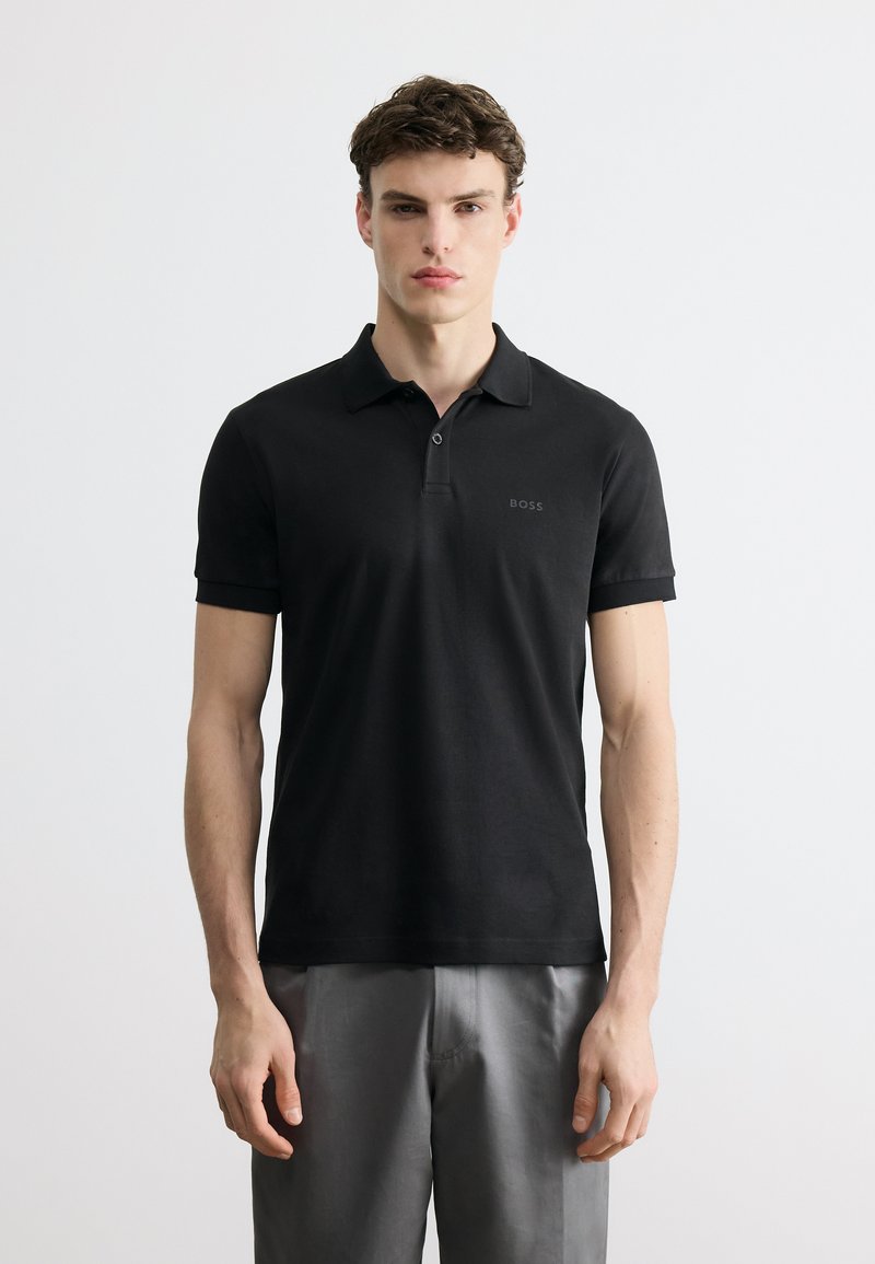 Νεαρός άνδρας που φοράει μαύρο πόλο μπλουζάκι BOSS και γκρι παντελόνι, στέκεται μπροστά σε απλό λευκό φόντο, κοιτώντας ευθεία μπροστά.