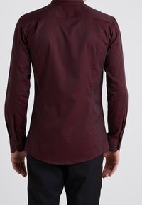 Camisa de manga larga, de color burdeos, con una textura suave, puños abotonados y un collar estándar, diseñada con un ajuste entallado en la parte posterior.