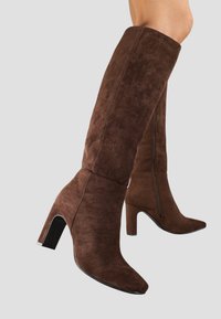 Bottes en daim marron jusqu'au genou avec un bout pointu et un talon épais. Détail de fermeture éclair sur le côté visible, texture douce et surface lisse.