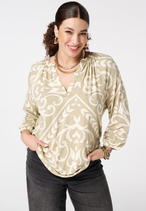 Lachende vrouw met krullend haar in een beige-wit blouse met patroon, zwarte broek, gouden oorringen, meerdere kettingen en armbanden.