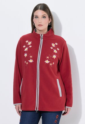 Femme portant une veste rouge à fermeture éclair avec une broderie florale blanche et un bord gris, debout devant un fond blanc uni.