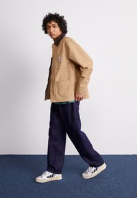 Veste en toile beige avec un col marron, poches avant et fermeture à boutons, associée à un jean bleu foncé et des baskets blanches sur un tapis bleu.
