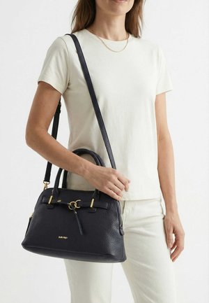 Mujer que lleva una camisa blanca de manga corta y pantalones, sosteniendo un bolso de cuero negro con herrajes en tono dorado y correa para el hombro.