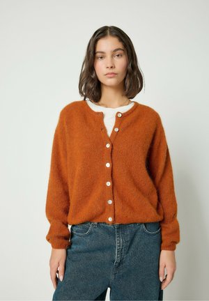 Kuldne, karvane kardigan ringi kaelusega, ees nööpidega ja pikkade varrukatega, koos kõrge vöökohaga siniste teksadega.