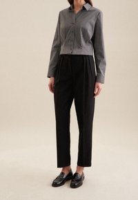 Person klädd i en grå långärmad knäppt skjorta, högmidjade svarta veckade byxor och svarta läder loafers mot en neutral bakgrund.