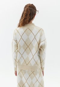 Krämfärgad tröja med beige och vit argyle-mönster, ribbad krage och nedkant, med en mjuk textur och avslappnad passform. Bakåtsikt visas.