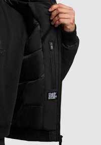 Innsiden av svart polstret jakke som viser quiltet tekstur, glidelåslomme og etikett. Har mykt fôr og ribbestrikkede mansjetter for komfort.