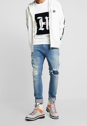 Sudadera blanca con capucha y logo, camiseta gráfica en blanco y negro, vaqueros desgastados de color azul claro y botas de senderismo multicolor con suelas negras.