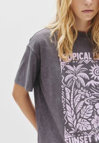 Grå oversized t-shirt med et tropisk solnedgangsdesign i lys pink med palmer og huse, lavet af blødt, tekstureret stof.